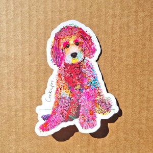 May include: A colourful sticker of a pink and white Cockapoo dog with a white background. The dog is sitting with its front paws crossed and its head tilted slightly to the side. The sticker has the word "Cockapoo" written in cursive on the left side.