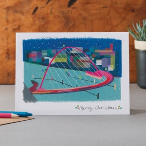 Può includere: Un biglietto di Natale con un'illustrazione di un ponte con un arco rosa e cavi bianchi. Lo sfondo raffigura un paesaggio urbano sotto un cielo blu stellato. Le parole "Merry Christmas" sono scritte in basso.
