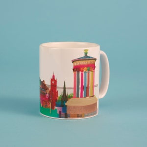 May include: White ceramic mug featuring a colourful illustration of a city skyline. The skyline includes a tall building with a clock tower, a monument with columns, and other buildings.
