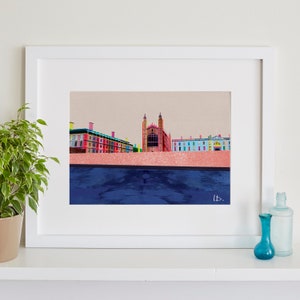 Op de afbeelding: Een ingelijste print van een kleurrijke illustratie van een gebouw met een reflectie van het gebouw in een waterpartij. Het gebouw is een mix van rood, geel, blauw en groen. De reflectie is donkerblauw. De print is ingelijst in een wit frame.