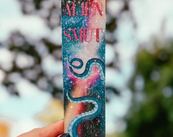 May include: A rectangular bookmark with a space-themed design. The top of the bookmark has the words "ALIEN SMUT" in pink. Below the text is a blue and pink galaxy design with a blue tentacle. The bookmark is held by a person.
