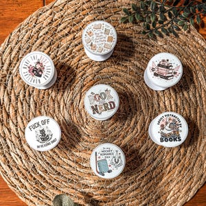 Puede incluir: Una colección de soportes blancos para teléfonos con varios diseños y frases temáticas de libros. Los soportes presentan ilustraciones de libros, flores y textos como "Book Nerd" y "Easily Distracted by Books". Están dispuestos sobre un mantel individual redondo y tejido.