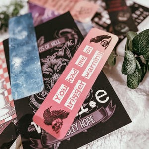 May include: A collection of decorative bookmarks with various designs and text. One pink bookmark reads "You had me at trigger warning" with raven and skull illustrations. Other bookmarks feature celestial and gothic themes.