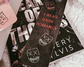 May include: A black bookmark with white and red text that reads "I READ DARK ROMANCE" and features three stylized mask designs. The bookmark is placed on top of a book with the title "HEART OF THORNS".