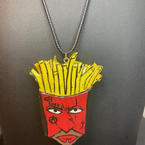 May include: A handmade pendant necklace featuring a cartoon-style design of a red and yellow french fry container with a face. The pendant hangs from a black cord. The container has the text "CD Lock" and other symbols.