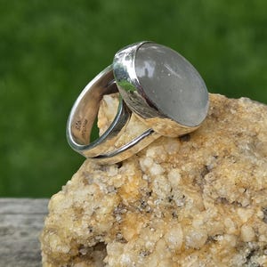 Puede incluir: Un anillo de plata con una gema grande, ovalada y gris pálido. El anillo está colocado sobre una roca texturizada de color marrón claro, sobre un fondo verde borroso. La banda del anillo está marcada con "925" y "michael".