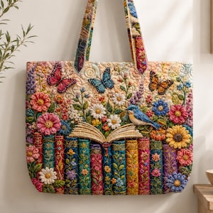 Op de afbeelding: Een gewatteerde tote bag met een levendig, veelkleurig ontwerp van een rij kleurrijke boeken met een open boek erbovenop. Het tafereel is versierd met diverse bloemen, een kleine blauwe vogel en drie vlinders in de kleuren roze, blauw en oranje.