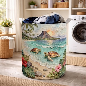 Könnte beinhalten: Ein runder Wäschekorb mit einer tropischen Strandszene. Das Design zeigt Palmen, einen Vulkan und zwei Meeresschildkröten im Ozean. Der Korb ist mit Kleidung gefüllt und hat Griffe.