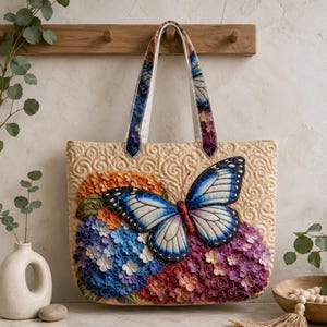Puede incluir: Un bolso tote acolchado de color beige con una gran mariposa bordada en azul y blanco sobre un racimo de flores de hortensia naranjas, azules y moradas. El bolso tiene dos asas de tela a juego con un estampado de mariposas azul y blanco.