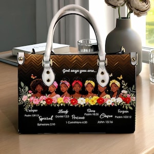 Puede incluir: Bolso de mano negro con asas blancas y herrajes dorados. El bolso presenta un panel decorativo con ilustraciones de mujeres y detalles florales. El texto en el bolso incluye "Dios dice que eres..." y varias palabras y versículos bíblicos inspiradores.