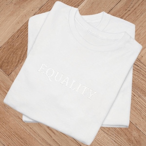 Könnte beinhalten: Zwei weiße T-Shirts sind ordentlich gefaltet und auf einer Holzoberfläche gestapelt. Das Wort "EQUALITY" ist weiß auf die Vorderseite des oberen Shirts gestickt. Die Shirts haben einen klassischen Rundhalsausschnitt.