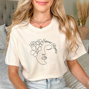 Könnte beinhalten: Cremefarbenes T-Shirt mit einer minimalistischen Strichzeichnung eines Gesichts, das mit Blumen verziert ist, begleitet von den Worten "Equal Rights" in einem stilvollen Schriftzug. Das Design ist in Schwarz gehalten. Das Shirt besteht aus einem weichen Material.