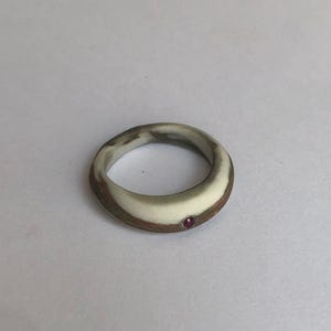 May include: A handcrafted ring with a unique design. The ring features a marbled effect with brown, white, and grey tones. A small, red gemstone is set into the band. The ring is a piece of jewellery.
