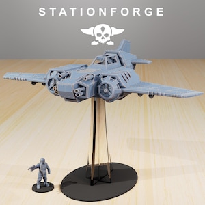 Può includere: Modello di aereo grigio chiaro con testo STATIONFORGE e logo teschio. Il modello è montato su un supporto con una piccola figura davanti. L'aereo ha ali, eliche e più canne di fucile.