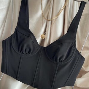 May include: A black bustier top with a structured bodice and thin straps. The top is displayed on a hanger with a gold layered necklace. The fabric has a slight sheen, and the background is a neutral color.