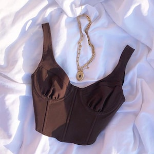 May include: A brown bustier top with wide straps and a structured bodice. The top is paired with a gold chain necklace with a pendant. The items are displayed on a white, wrinkled fabric.