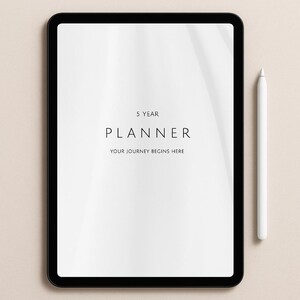 May include: A black tablet with a white screen displaying the text "5 YEAR PLANNER YOUR JOURNEY BEGINS HERE." A white stylus rests beside the tablet on a neutral background. The image is clean and minimalist.