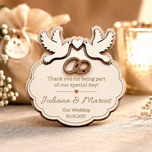 May include: Wooden wedding favour featuring two doves, rings, and text. The text reads "Thank you for being part of our special day!" and the names "Juliana & Marcos" with the wedding date "30.05.2027".