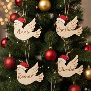 Könnte beinhalten: Weihnachtsbaumschmuck in Form von Tauben, jede mit Weihnachtsmütze und Namen: Anna, Cynthia, Roman und Charlie. Die Ornamente sind aus hellem Holz gefertigt und hängen zwischen roten und goldenen Kugeln.
