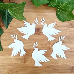 Puede incluir: Cinco recortes de palomas blancas, cada una sosteniendo una pequeña rama de olivo en sus picos. Las palomas están dispuestas sobre una superficie de madera, con hojas verdes de hiedra en el fondo. Las palomas tienen las alas extendidas, símbolo de paz.