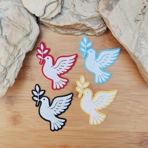 Puede incluir: Cuatro recortes de palomas coloridas, cada una sosteniendo una rama de olivo. Las palomas son blancas con detalles en rojo, azul, negro y amarillo. Sobre una superficie de madera, con fondo neutro.