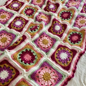 May include: A handmade crochet blanket composed of numerous square motifs featuring a floral-inspired design. The squares are joined with white yarn and display a vibrant color palette of magenta, light pink, mustard yellow, and sage green.