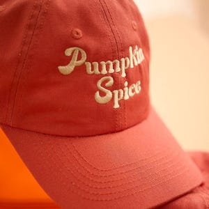 Puede incluir: Gorra de béisbol roja con la inscripción "Pumpkin Spice" bordada en crema. La gorra tiene una visera curva y un diseño clásico de seis paneles, ideal para el uso diario.