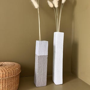 May include: Two tall, rectangular vases with textured surfaces. One is gray and white, the other is white. Each holds a bundle of fluffy, cream-colored dried flowers. The vases are set against a muted green wall.