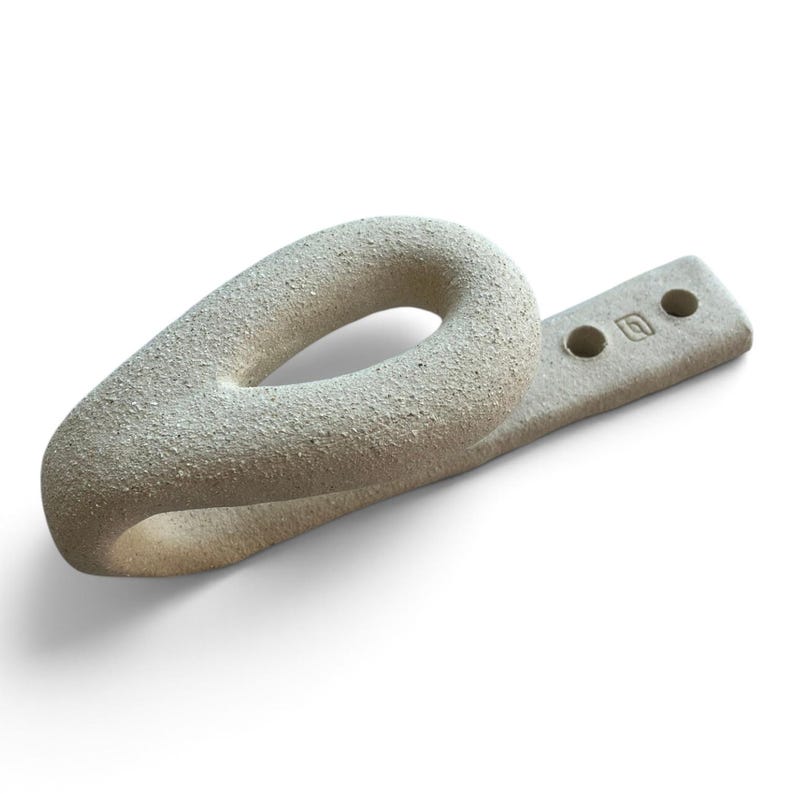 May include: A beige, textured ceramic wall hook with a teardrop-shaped loop and a flat, rectangular mounting base. The base features two circular holes for installation and a small, square maker's mark stamped into the surface of the clay.