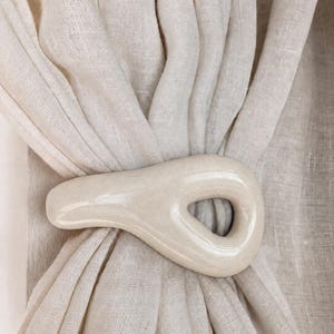 May include: A cream-colored, smooth, ceramic curtain tieback with an organic, asymmetrical, teardrop-like shape and a central opening, holding together a gathered, light-colored, semi-sheer linen-textured fabric curtain panel.