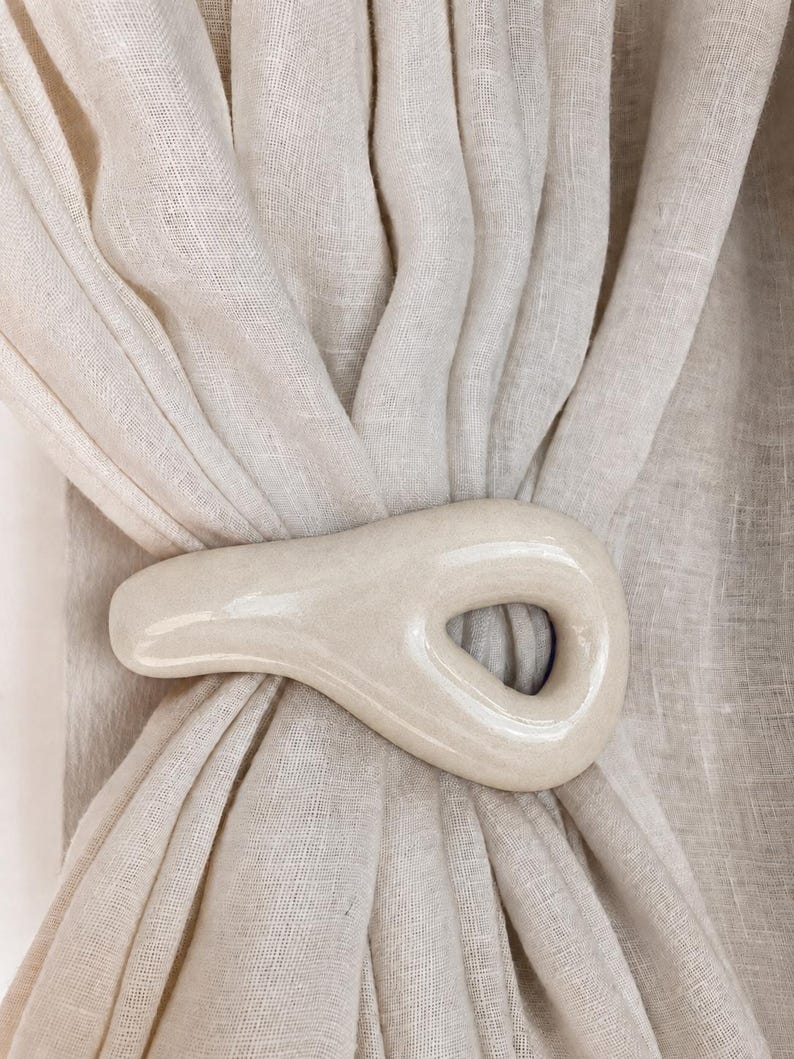 May include: A cream-colored, smooth, ceramic curtain tieback with an organic, asymmetrical, teardrop-like shape and a central opening, holding together a gathered, light-colored, semi-sheer linen-textured fabric curtain panel.