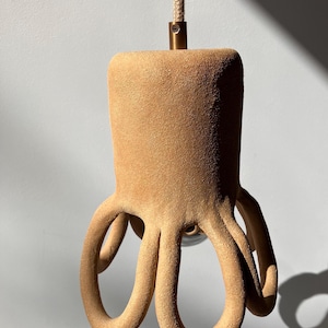 Peut inclure: Suspension luminaire en forme de poulpe de couleur beige. Le luminaire présente un sommet cylindrique et huit tentacules incurvés. Il est suspendu à un cordon beige et à un raccord en laiton. Décoration intérieure.