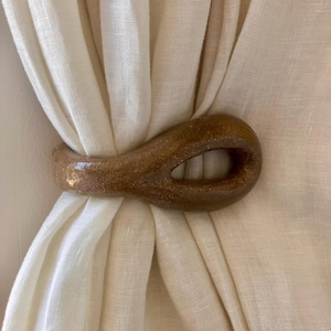 May include: A smooth, brown, teardrop-shaped ceramic curtain tieback with a glossy, speckled finish. The object features a central open loop and is wrapped around a piece of off-white, semi-sheer fabric, holding the material together in a gathered fold.