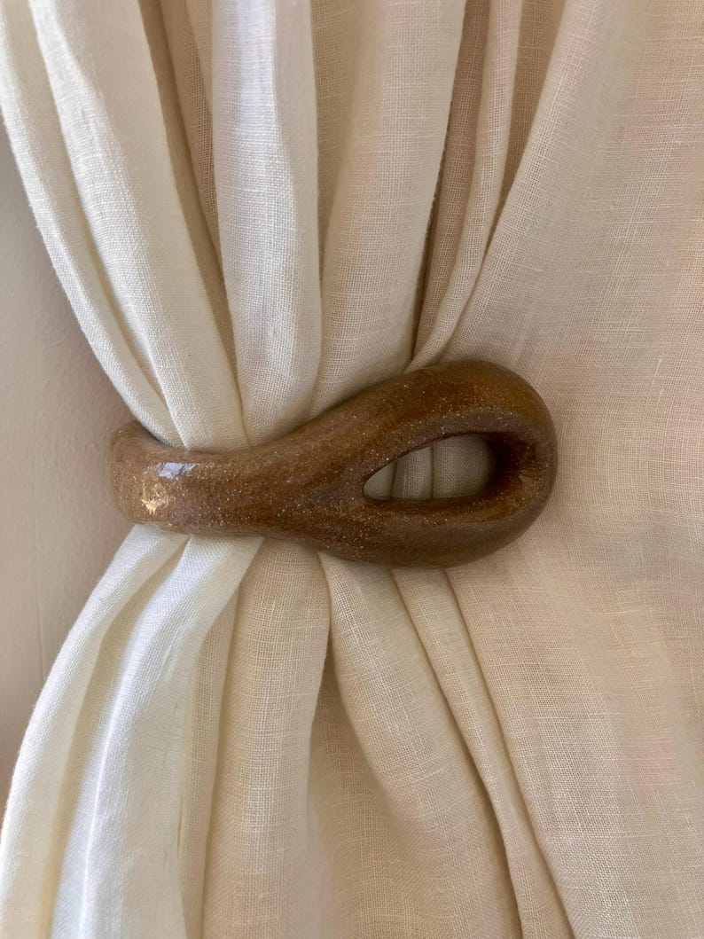 May include: A smooth, brown, teardrop-shaped ceramic curtain tieback with a glossy, speckled finish. The object features a central open loop and is wrapped around a piece of off-white, semi-sheer fabric, holding the material together in a gathered fold.