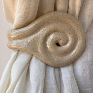 May include: A beige ceramic curtain tieback with a glossy finish and a speckled texture, shaped into a spiral design. The object is secured around a piece of light-colored, semi-sheer fabric, holding the material in a gathered, draped position.