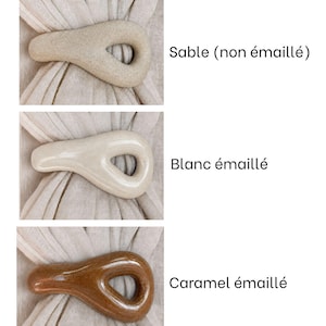 May include: Three teardrop-shaped ceramic napkin rings shown in different finishes. The top is matte beige, the middle is glossy off-white, and the bottom is glossy caramel brown. Each ring is displayed over a light beige fabric napkin.