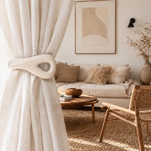May include: A smooth, off-white ceramic curtain tieback with an elongated, teardrop-shaped loop design. The object is secured around a sheer, light-colored fabric curtain, holding the material in a gathered, draped position with its glossy finish.