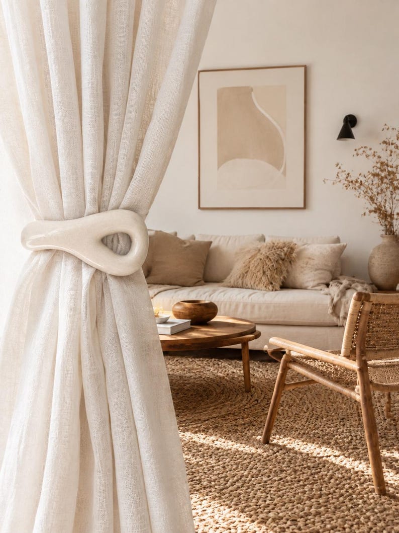 May include: A smooth, off-white ceramic curtain tieback with an elongated, teardrop-shaped loop design. The object is secured around a sheer, light-colored fabric curtain, holding the material in a gathered, draped position with its glossy finish.