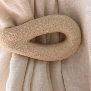 May include: A beige, textured, oval-shaped curtain tieback with a central cutout, securing a piece of off-white, semi-sheer fabric. The tieback has a granular, stone-like surface finish and is positioned horizontally across the gathered textile.