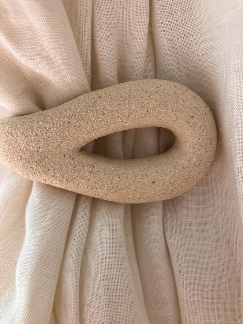 May include: A beige, textured, oval-shaped curtain tieback with a central cutout, securing a piece of off-white, semi-sheer fabric. The tieback has a granular, stone-like surface finish and is positioned horizontally across the gathered textile.