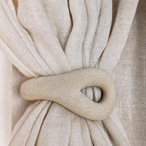 May include: A beige, textured, teardrop-shaped curtain tieback holding together a gathered, semi-sheer, light-colored fabric curtain. The tieback has a smooth, stone-like appearance with a central opening and a matte, speckled finish.