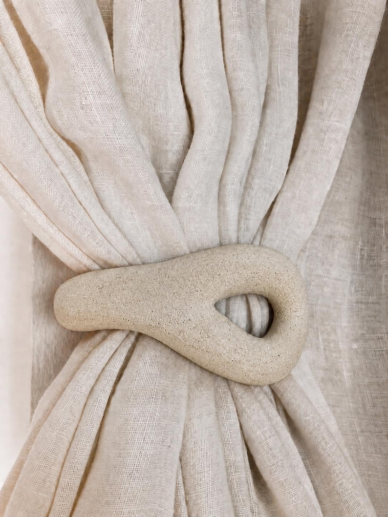 May include: A beige, textured, teardrop-shaped curtain tieback holding together a gathered, semi-sheer, light-colored fabric curtain. The tieback has a smooth, stone-like appearance with a central opening and a matte, speckled finish.