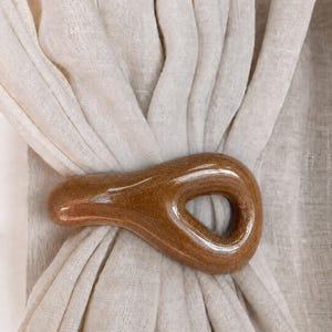 May include: A smooth, glossy, brown ceramic curtain tieback with a teardrop-shaped opening, securing a piece of off-white, semi-sheer linen fabric. The ceramic piece has a speckled, earthy texture and a reflective finish that catches the light.