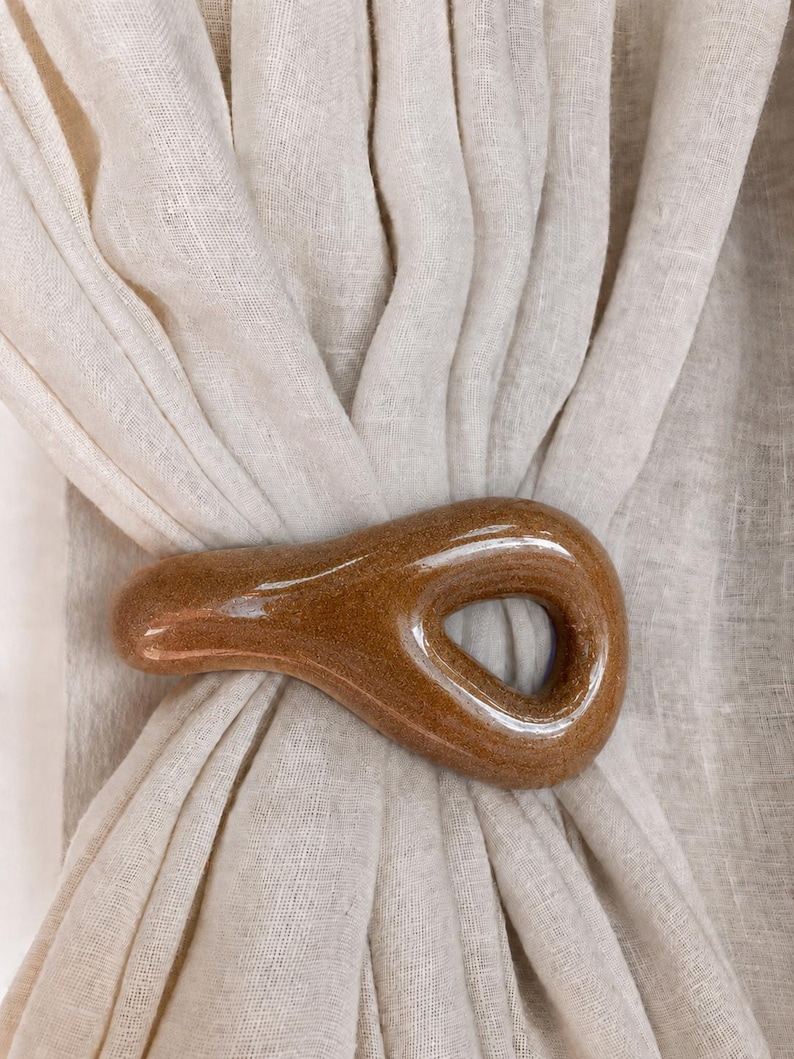 May include: A smooth, glossy, brown ceramic curtain tieback with a teardrop-shaped opening, securing a piece of off-white, semi-sheer linen fabric. The ceramic piece has a speckled, earthy texture and a reflective finish that catches the light.