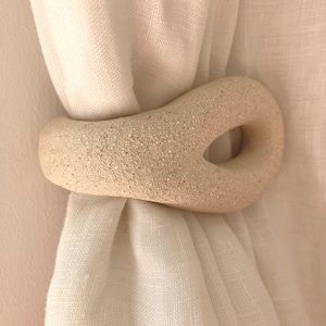 May include: A beige, textured, organic-shaped ceramic curtain tieback holding a gathered white fabric curtain. The tieback features a smooth, speckled surface with a central open loop, providing a functional way to secure the curtain in place.