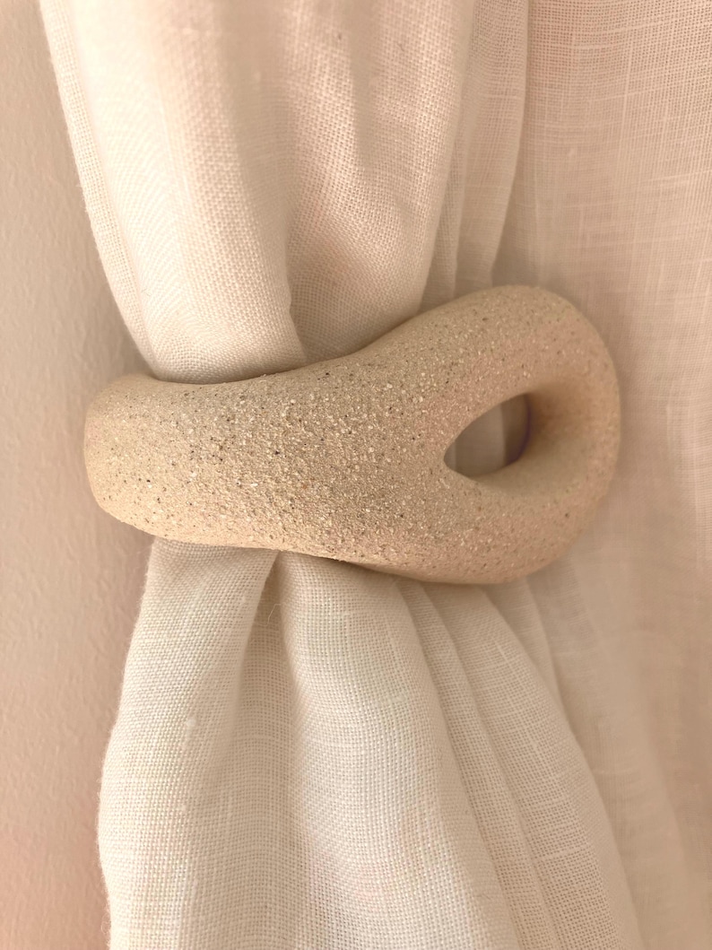 May include: A beige, textured, organic-shaped ceramic curtain tieback holding a gathered white fabric curtain. The tieback features a smooth, speckled surface with a central open loop, providing a functional way to secure the curtain in place.