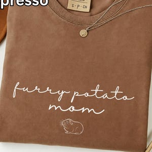 May include: Espresso-colored t-shirt with the text "furry potato mom" and a guinea pig illustration. The shirt has a crew neck and a Comfort Colors label. A silver necklace with a small pendant is draped across the shirt.
