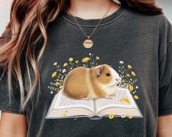 May include: Dark grey t-shirt featuring a guinea pig sitting on an open book. The guinea pig is brown and white, surrounded by yellow flowers. The book has text on the pages. A casual, comfortable tee.