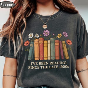 May include: A charcoal gray short-sleeve t-shirt featuring a graphic of a row of colorful books with flowers emerging from the tops. The text "I'VE BEEN READING SINCE THE LATE 1900s" is printed in a cream-colored font below the illustrated books.