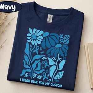 May include: Navy blue t-shirt with a floral design in shades of blue, including flowers and ribbons. The text "I wear blue for my custom" is printed at the bottom. The shirt is folded on a light-colored surface.